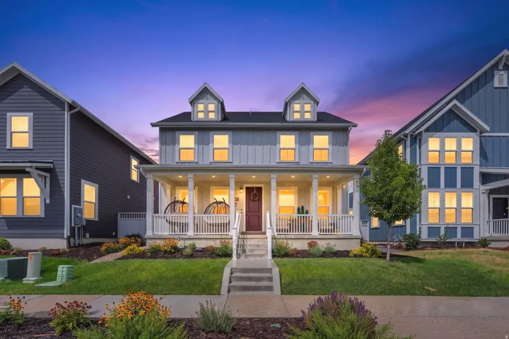 Classic Colonial-style residence featuring a symmetrical facade with dual peaked dormers, expansive front porch with turned balusters and columns, and warm exterior lighting throughout
