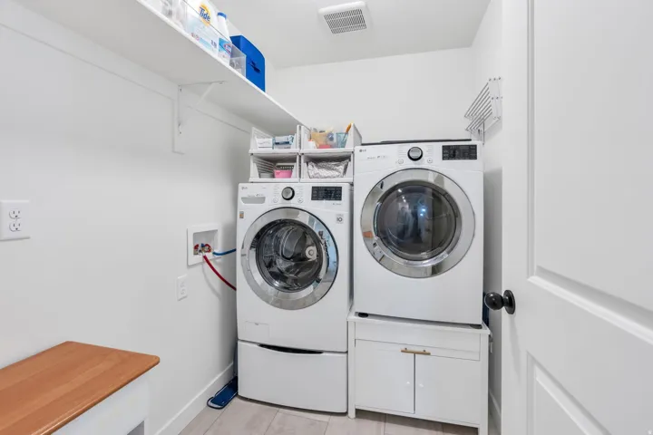 Modern laundry room featuring side-by-side front-load washer and dryer with pedestals, stainless steel appliances, white cabinetry with quartz countertop workspace, floating shelving for storage, and neutral finishes throughout