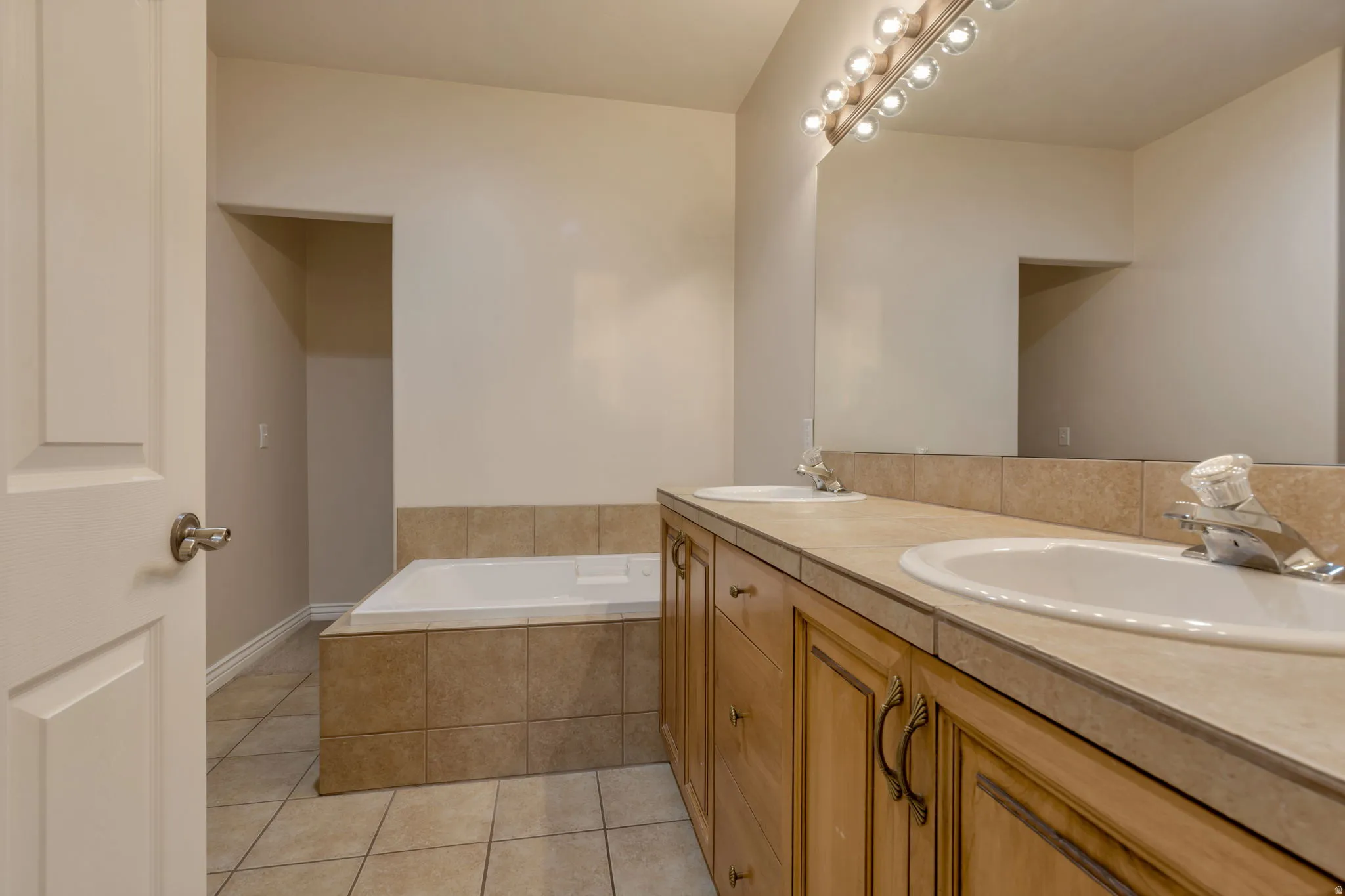 Dual undermount sinks with chrome faucets, wood-finish cabinetry with decorative pulls, integrated vanity mirror, built-in soaking tub, and tile flooring