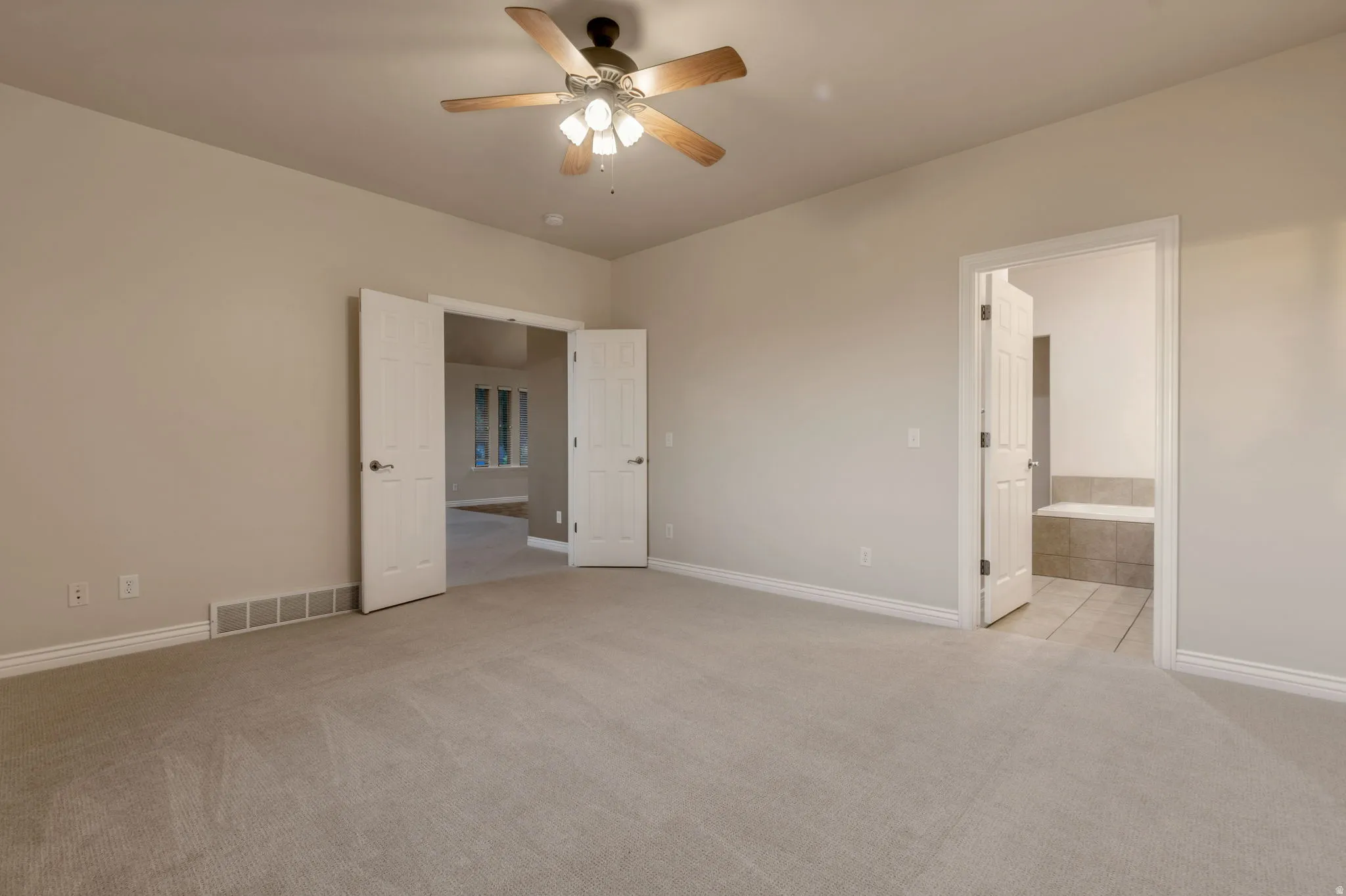 Ceiling fan with light fixture, neutral carpet flooring, white trim, double entry doors, and an en-suite bathroom with a built-in tub