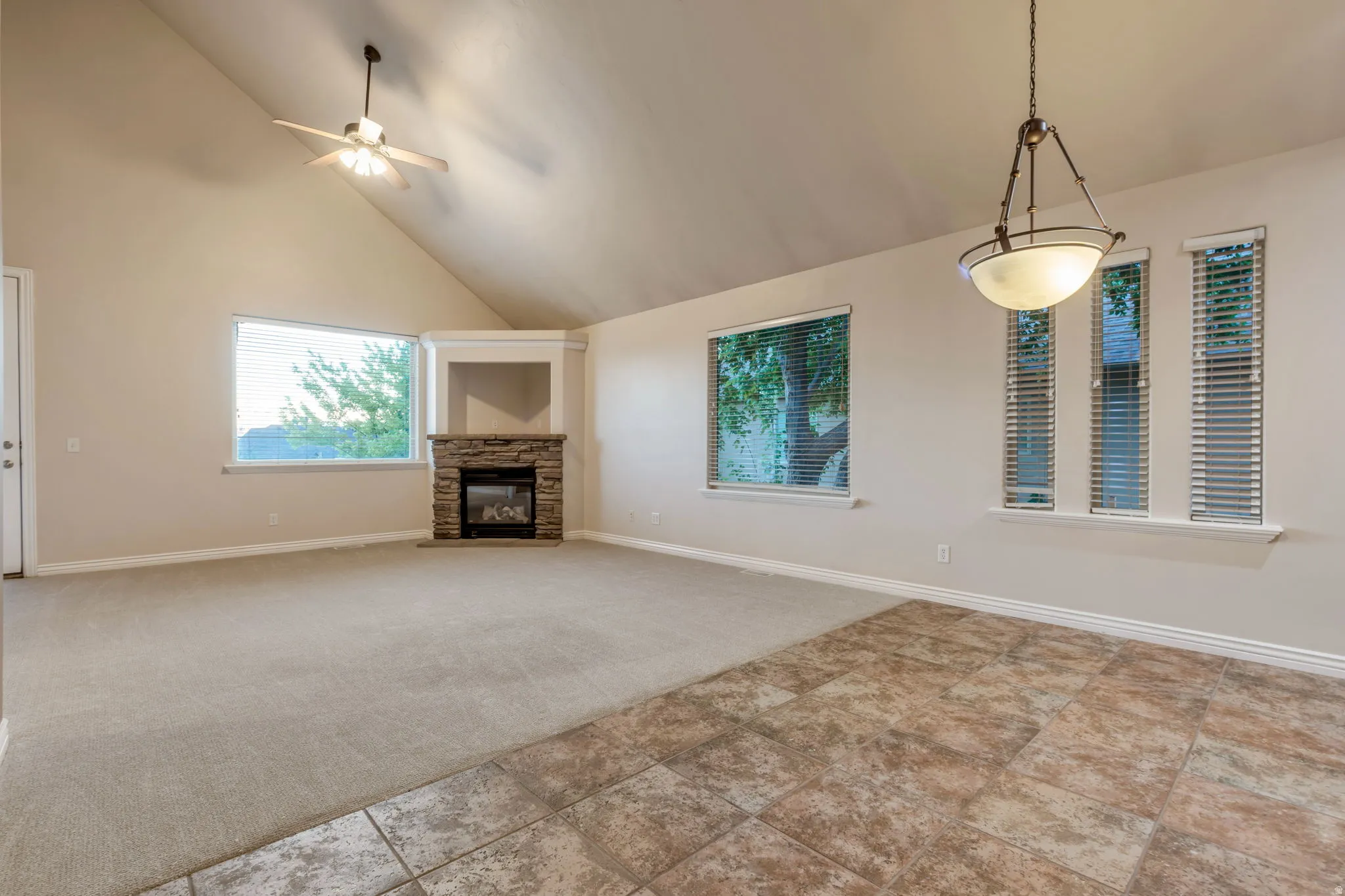 Vaulted ceiling with an attached ceiling fan, stone-clad fireplace with built-in shelving, mixed flooring with carpet and tile, and multiple windows providing natural light