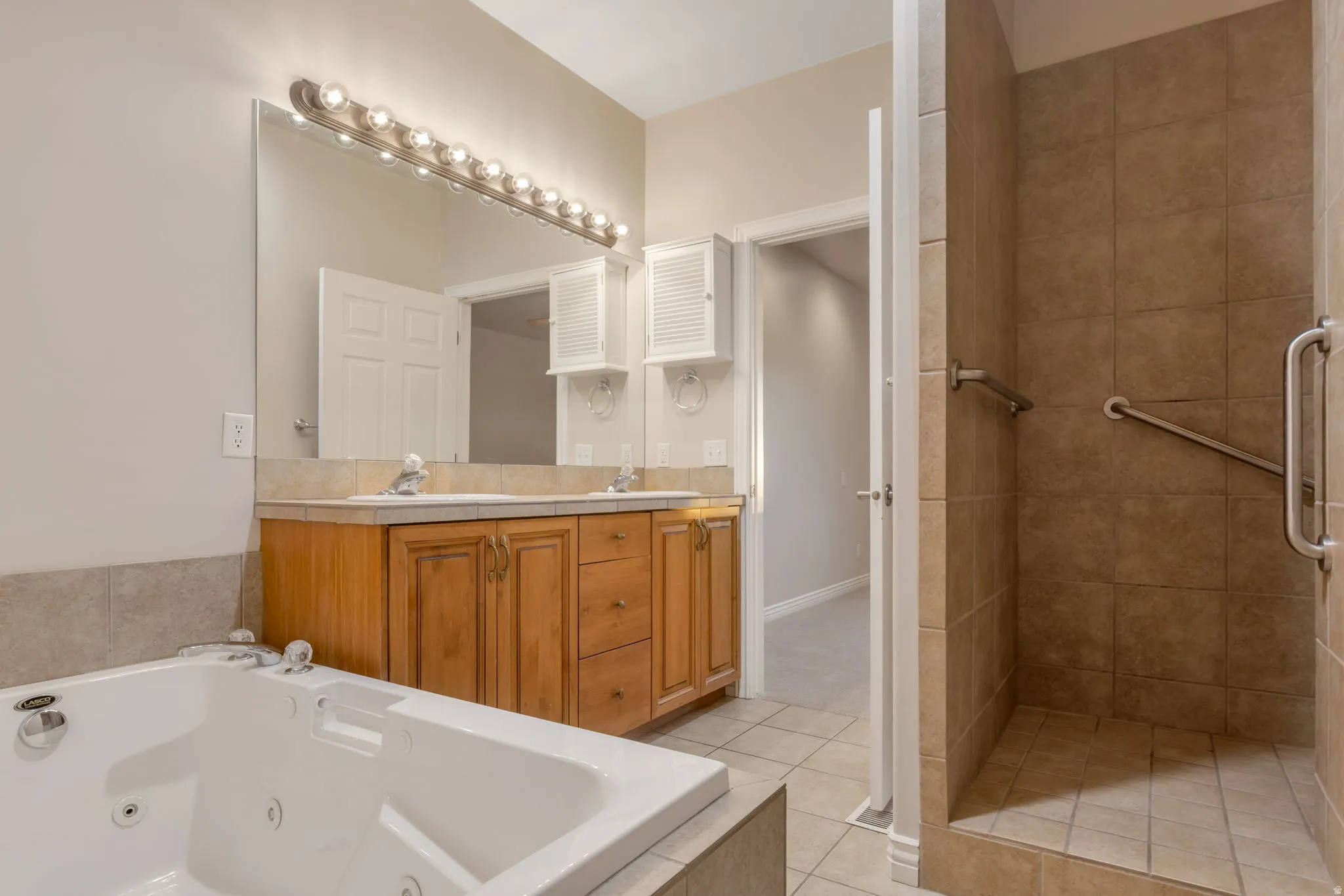 Dual vanity with wood-finish cabinetry, a large built-in mirror with strip lighting, a jetted soaking tub, and a tiled walk-in shower with grab bars