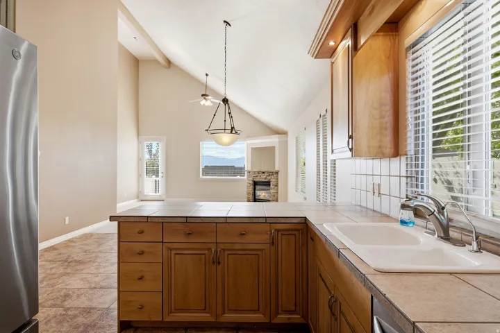 Vaulted ceilings, a stone-faced fireplace, a tiled kitchen island, wood-finish cabinetry, and a large window above the sink