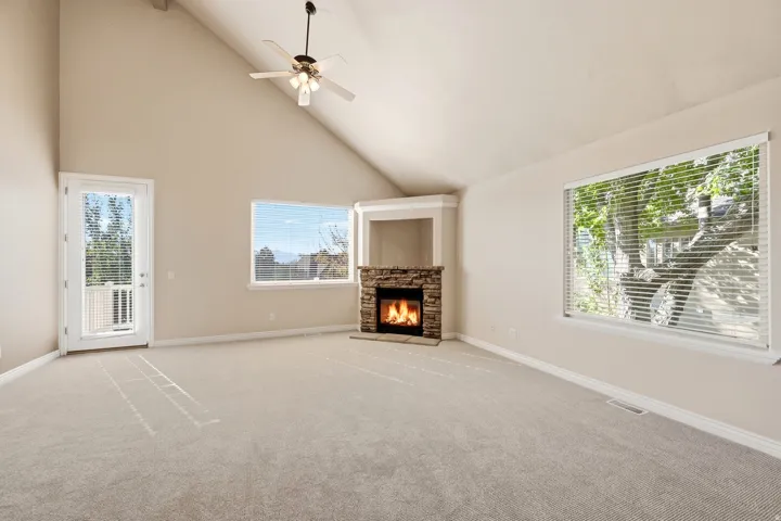 Vaulted ceiling with a ceiling fan, stone-clad fireplace with a recessed mantel, light-toned carpet flooring, multiple windows, and a glass-paneled door