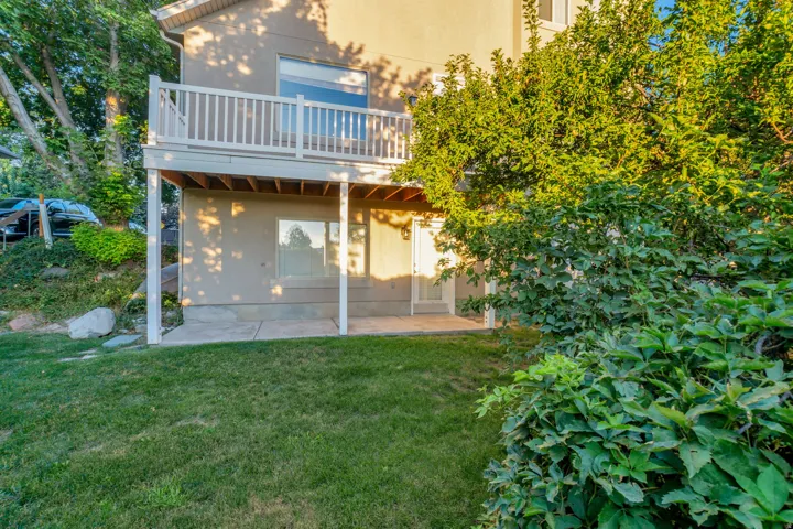 Rear facade with a second-story balcony featuring white railings, a ground-level patio, and surrounding mature landscaping