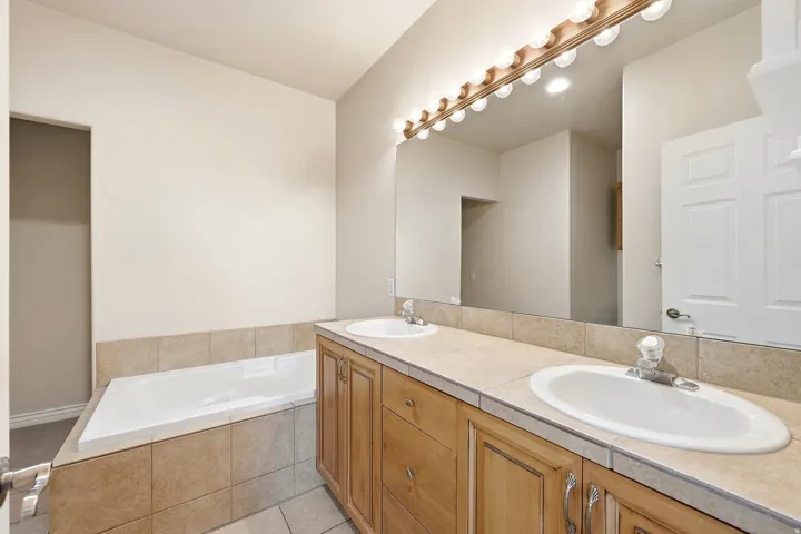Dual vanity with integrated sinks, wood-finish cabinetry, tile countertop, and a built-in bathtub with a tiled surround