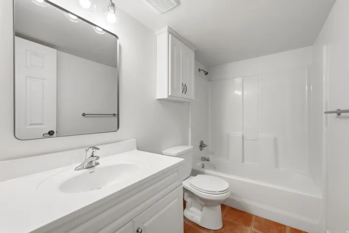 White vanity with an integrated sink and chrome faucet, large rectangular mirror, white wall-mounted storage cabinet, and a white tub with integrated shower surround