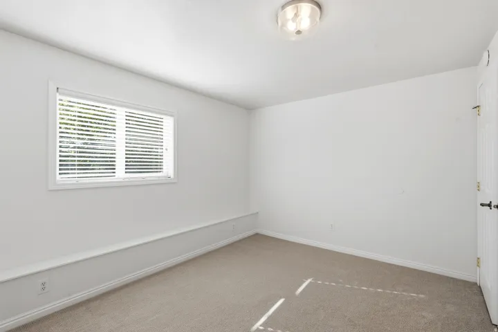 Neutral-toned carpeting paired with white walls, a single window with horizontal blinds, and a flush-mount ceiling light fixture