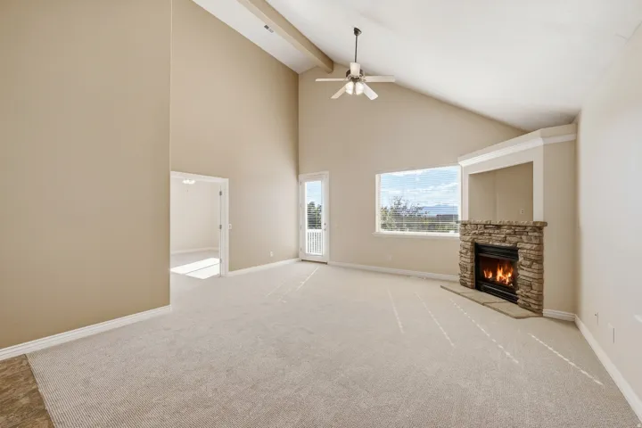 Vaulted ceilings, stone-faced fireplace, ceiling fan, large window, and light-toned carpeting