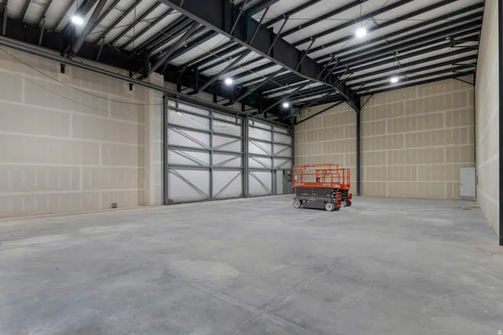 Concrete flooring, high ceilings with exposed metal trusses, industrial-style lighting fixtures, and large overhead door