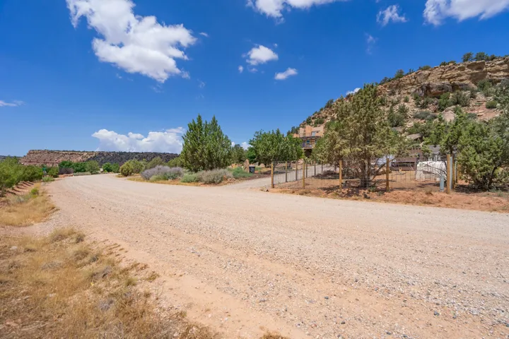 Gravel driveway leading to a fenced property with mature trees and a distant mesa landscape