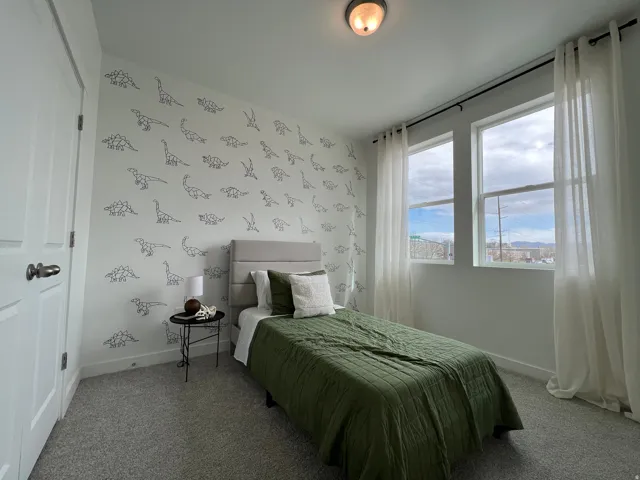 Bright secondary bedroom with large dual windows framing expansive city vistas, dinosaur-motif wallpaper, neutral carpet flooring, and a small side table
