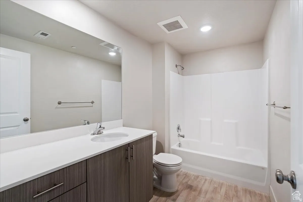 Modern bathroom featuring a wood-finish vanity with dual-sink white countertop, large frameless mirror with integrated lighting, shower enclosure with glass panel, ceramic toilet, and wood-finish flooring throughout