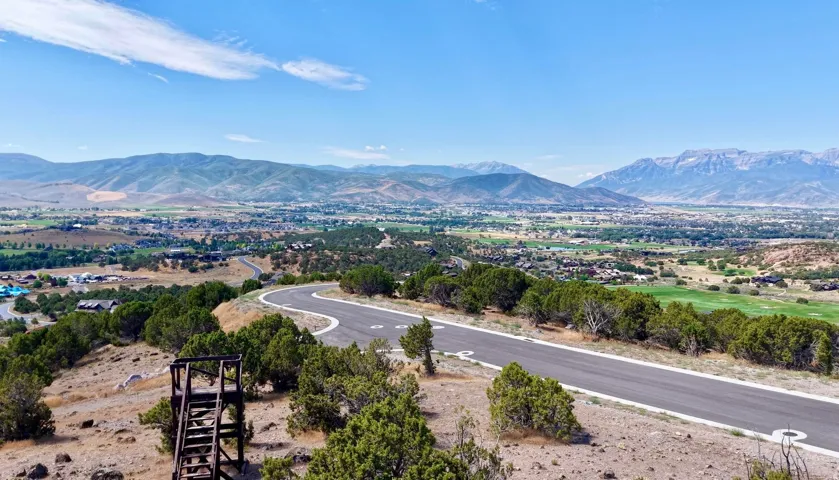 Winding paved road, surrounded by natural shrubbery, with distant mountain ranges and a developed valley