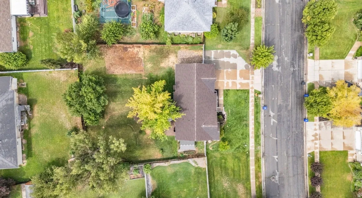 Aerial view showcasing a well-proportioned residential lot with mature landscaping, established trees throughout the grounds, manicured lawn areas, and a dark-roofed home situated on a tree-lined street in an established neighborhood