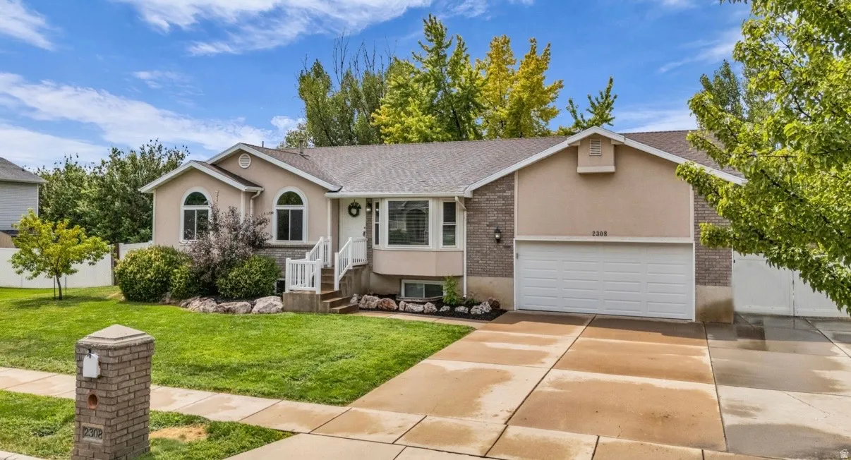 Split-level residence featuring a two-car garage, stucco and brick veneer exterior, arched windows, and a landscaped front yard with mature trees