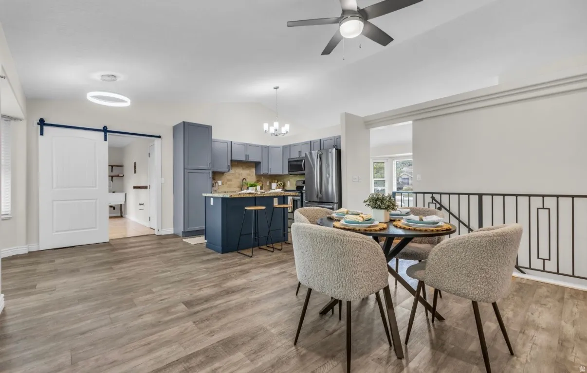 Open-concept living showcasing wood-finish flooring throughout, a chef's kitchen with navy cabinetry and stainless steel appliances, a dining area with round table seating, a ceiling fan with integrated lighting, and a sliding barn door entry