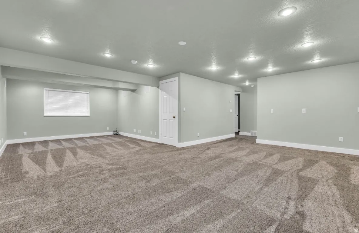Spacious basement recreation room featuring recessed ceiling lighting, neutral wall finishes, patterned carpet flooring, and multiple wall outlets