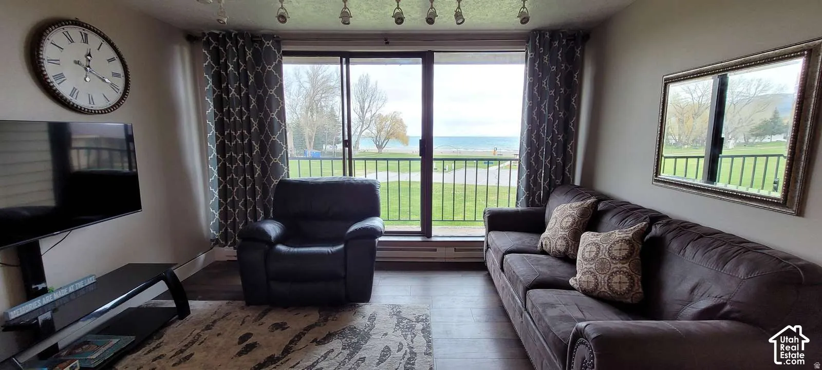Bright living room with expansive glass doors and windows framing scenic outdoor vistas, neutral wall finishes, wood-finish flooring, a wall-mounted clock, and ample natural light throughout the space