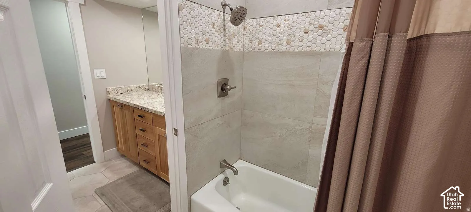 Bathtub and shower combination with hexagonal tile detailing, brushed nickel fixtures, a wood-finish vanity, and a granite-style countertop