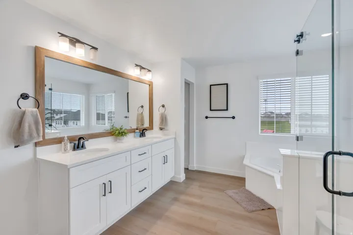 Dual vanity with white cabinetry and dark hardware, large wood-framed mirror, contemporary light fixtures, and glass-enclosed shower