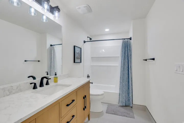 Basement bathroom with light wood-finish vanity with drawer storage, white stone-finish countertop, single basin sink, matte black fixtures, and a white tub and shower combination.
