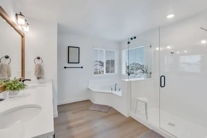 Dual vanity with white countertops, corner soaking tub, glass-enclosed shower, wood-finish flooring, and natural light