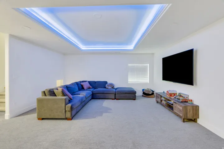 Recessed ceiling lighting, tray ceiling with cove lighting, wall-mounted television, window with blinds, and light gray carpet flooring
