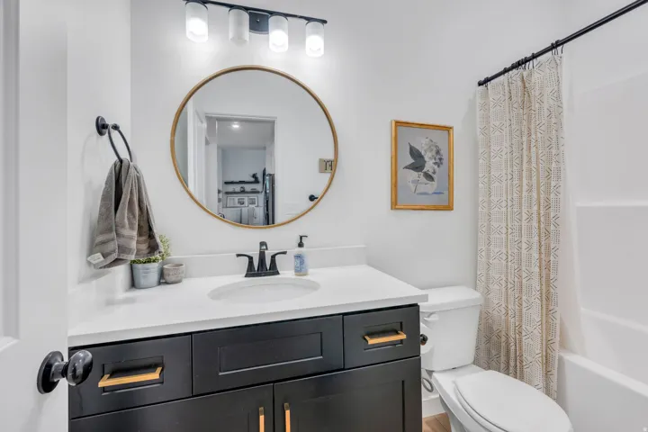 White rectangular bathtub with a shower rod, dark wood-finish vanity with gold-tone hardware, white countertop, and a round gold-tone framed mirror