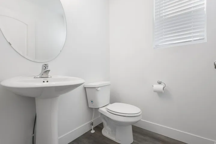 Pedestal sink with chrome faucet, circular wall mirror, wood-finish flooring, and window with horizontal blinds