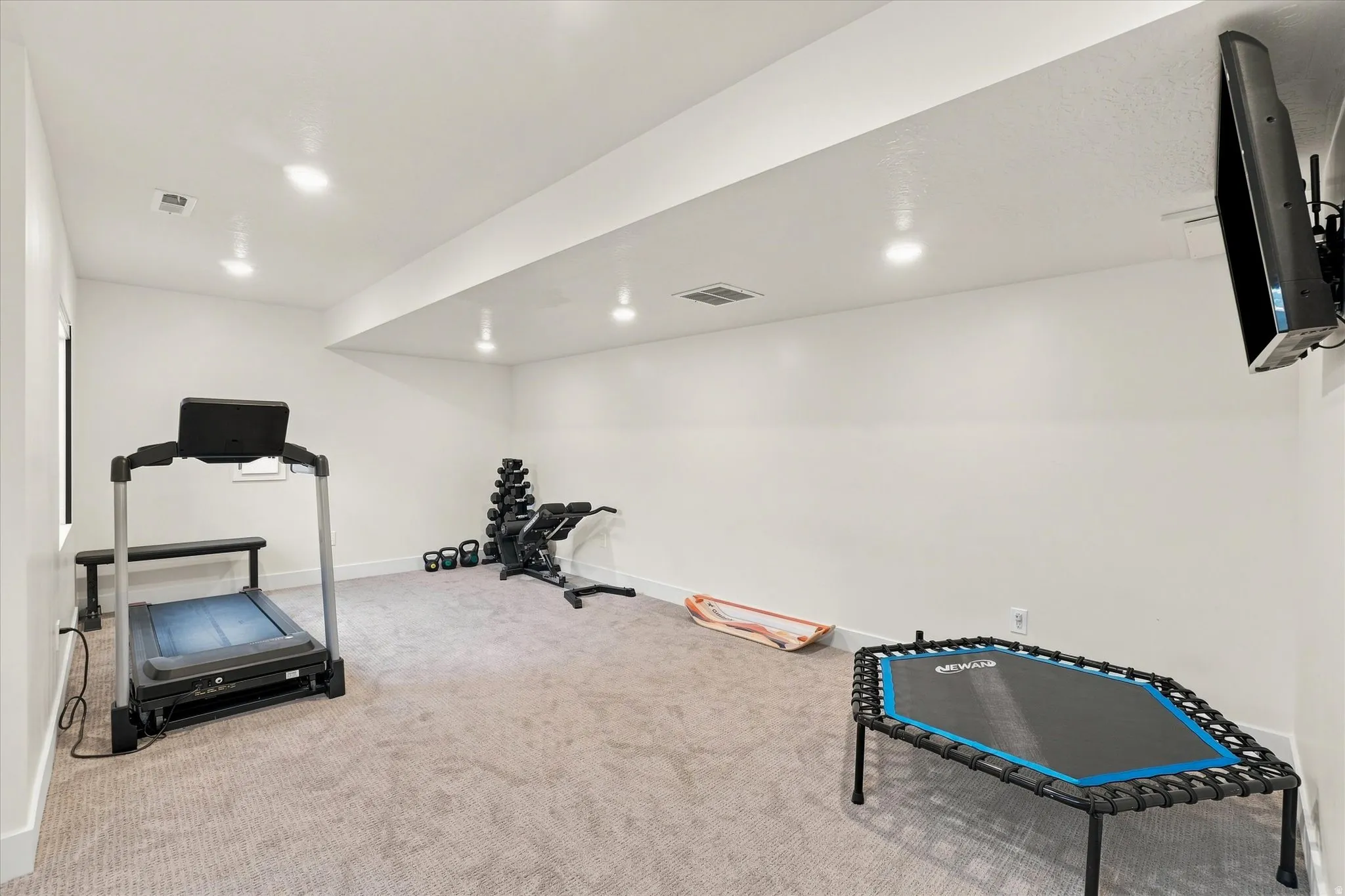 Recessed lighting, wall-mounted television bracket, neutral-toned carpeting, and a window providing natural light