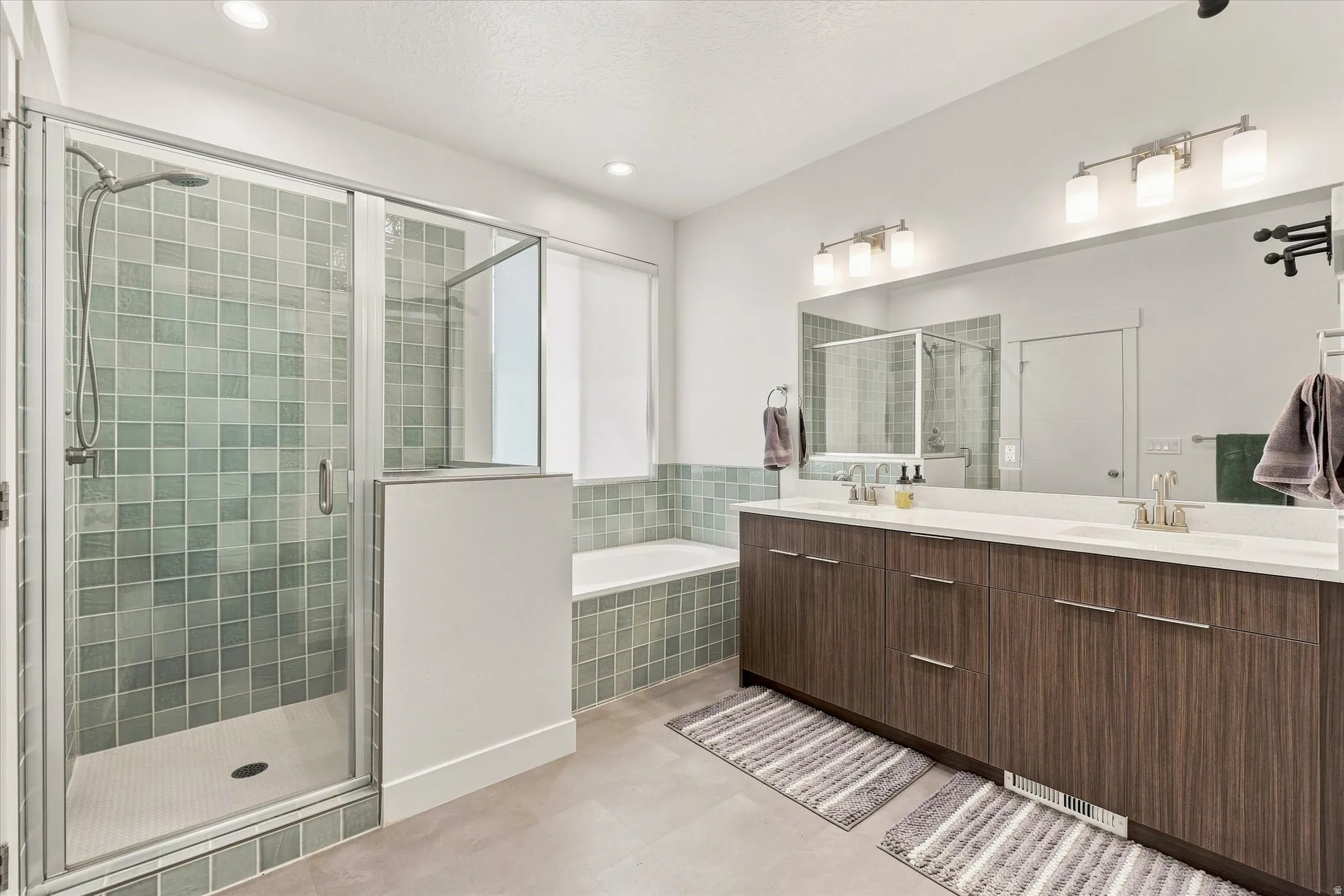 Walk-in shower with glass enclosure and mosaic tile, deep soaking tub, dual vanity with wood-finish cabinetry, and large mirrored wall