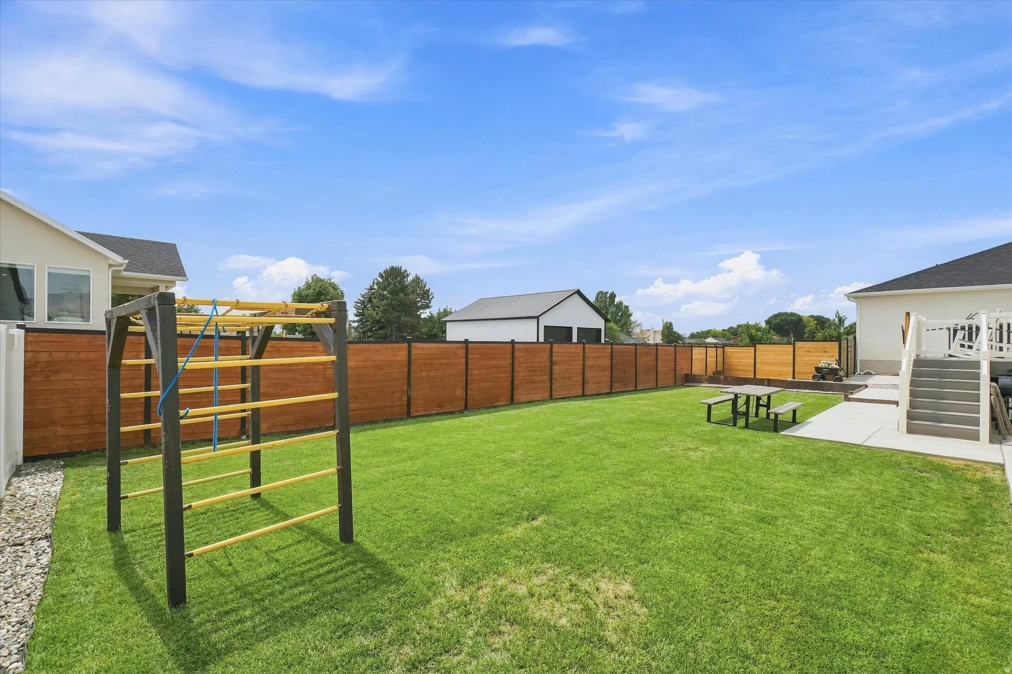 Expansive lawn area with a perimeter wood fence, concrete patio, and exterior stairs leading to an upper deck