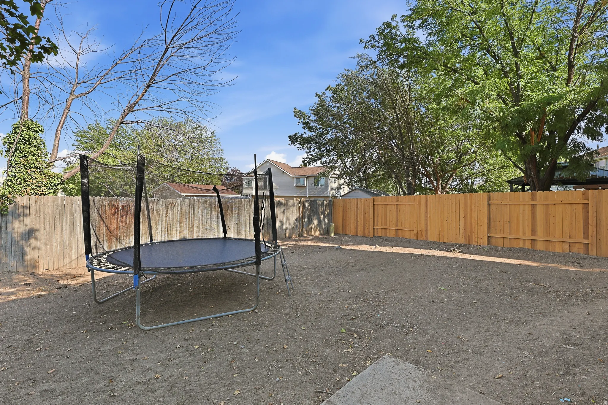 Spacious backyard oasis featuring a trampoline with safety enclosure, mature shade trees, and wooden privacy fencing