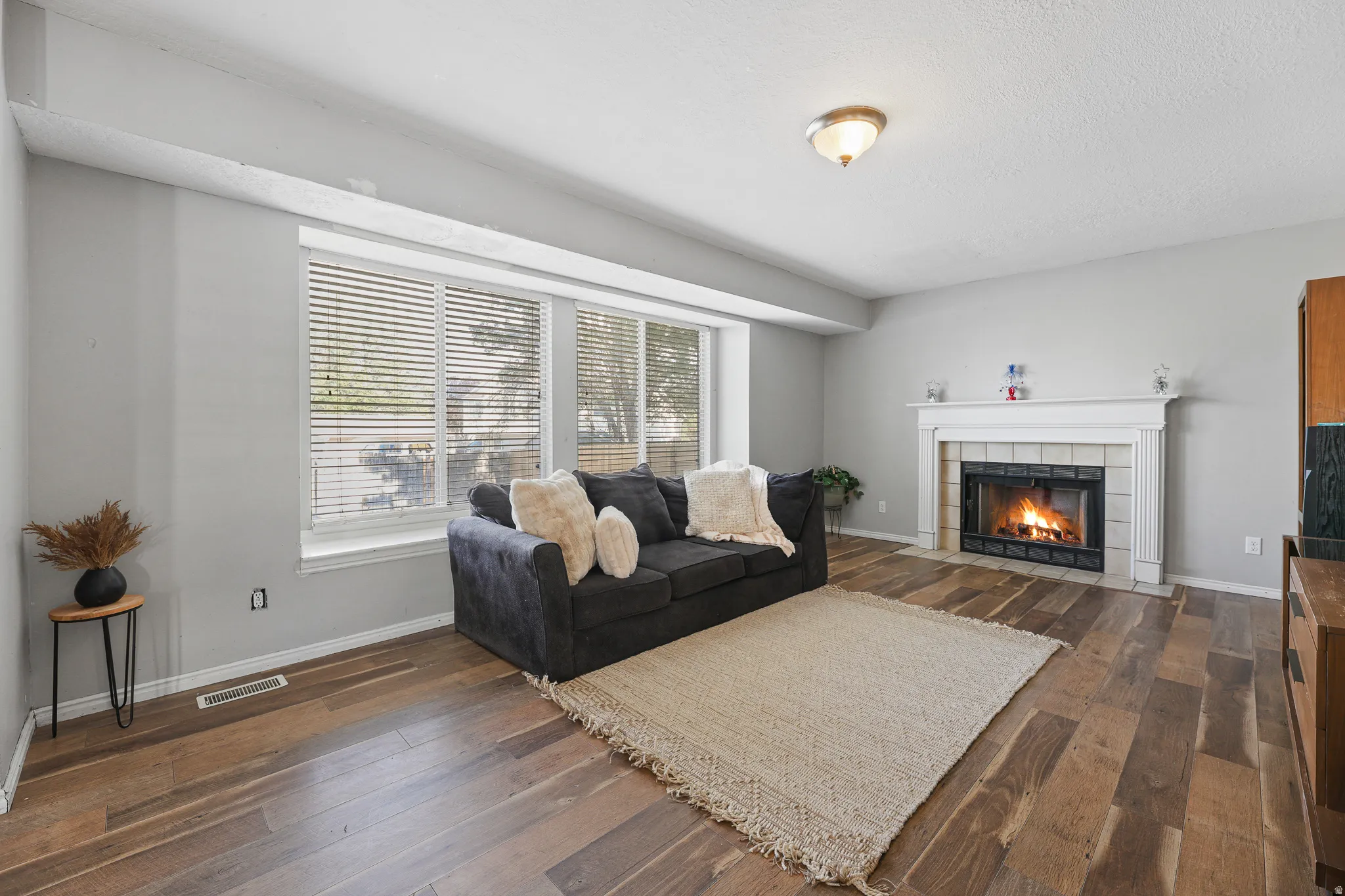 Spacious living area featuring wood-finish flooring, a large window with blinds, and a built-in fireplace with a white mantel and tile surround