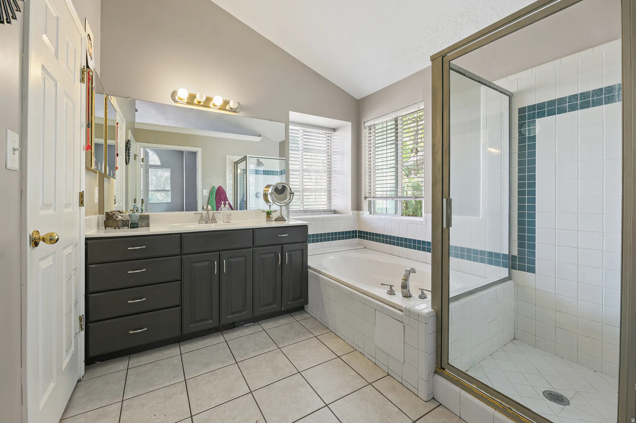 Dual-sink vanity with dark cabinetry, integrated soaking tub, separate glass-enclosed shower, tiled flooring, and vaulted ceiling