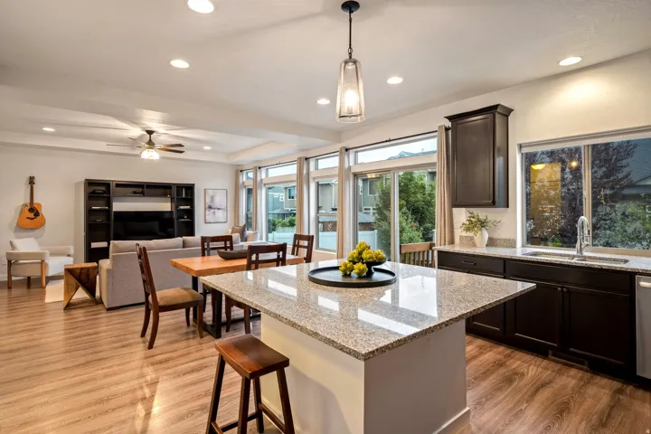 Open-concept kitchen and dining area featuring a large quartz-topped island with seating, stainless steel appliances, dark cabinetry with integrated hardware, recessed and pendant lighting, wood-finish flooring, and expansive multi-pane glass doors and wi