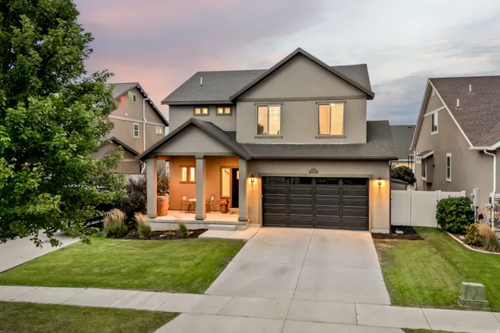 Modern two-story residence featuring a charcoal-finish garage door, illuminated entry portico with warm-toned lighting, manicured front lawn with mature landscaping, and a wide concrete driveway
