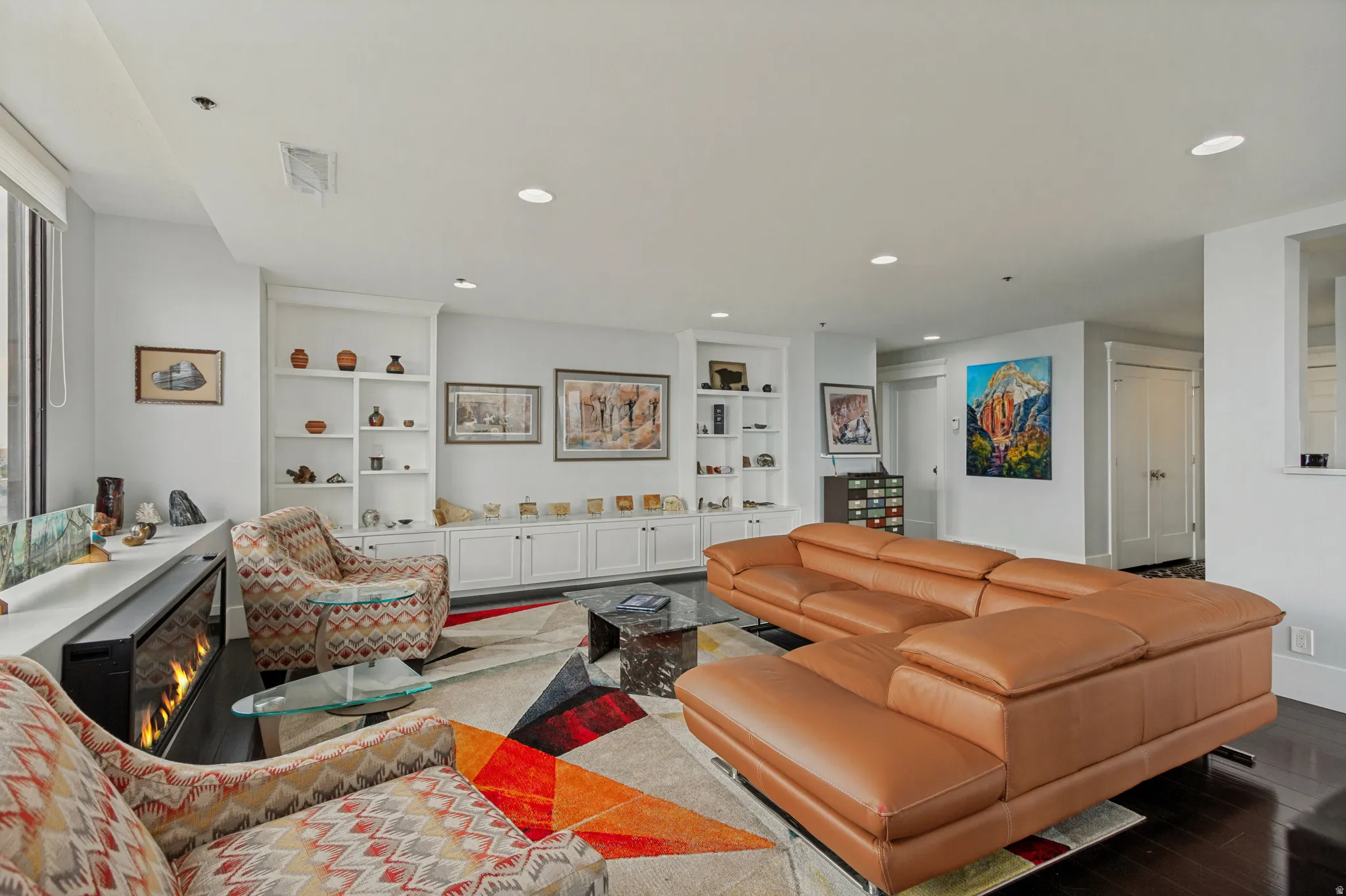 Open-concept living area featuring a modern caramel leather sectional sofa, recessed ceiling lighting, dark wood-finish flooring, and custom white built-in cabinetry with shelving throughout