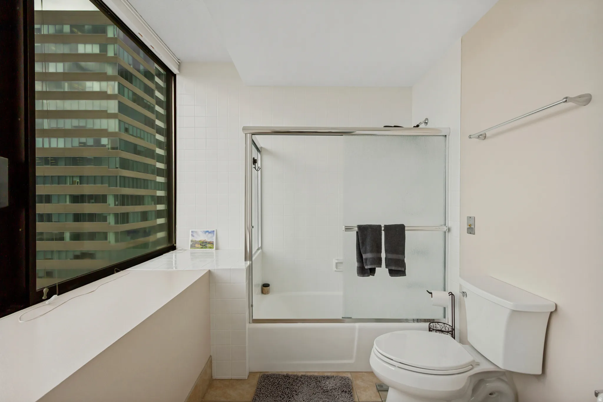 Large window with roller shade, white tiled shower, shower with sliding glass door, white bathtub, and toilet
