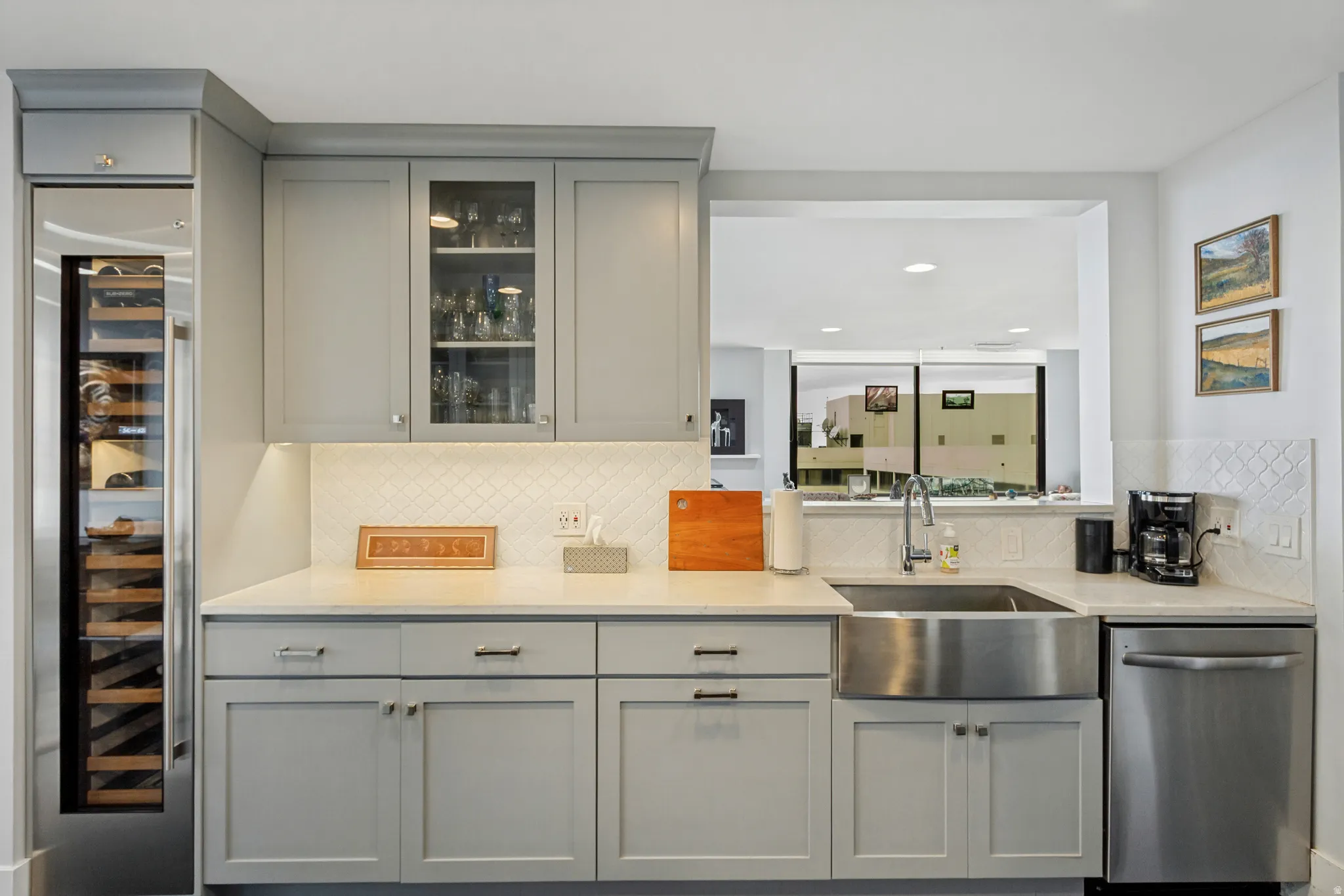 Built-in wine refrigerator, glass-front upper cabinetry, under-cabinet lighting, farmhouse-style stainless steel sink, and stainless steel dishwasher