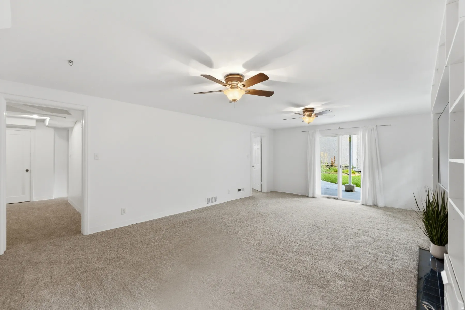 Spacious room featuring carpeting, two ceiling fans with integrated lighting, built-in shelving, and sliding glass doors to outdoor paving
