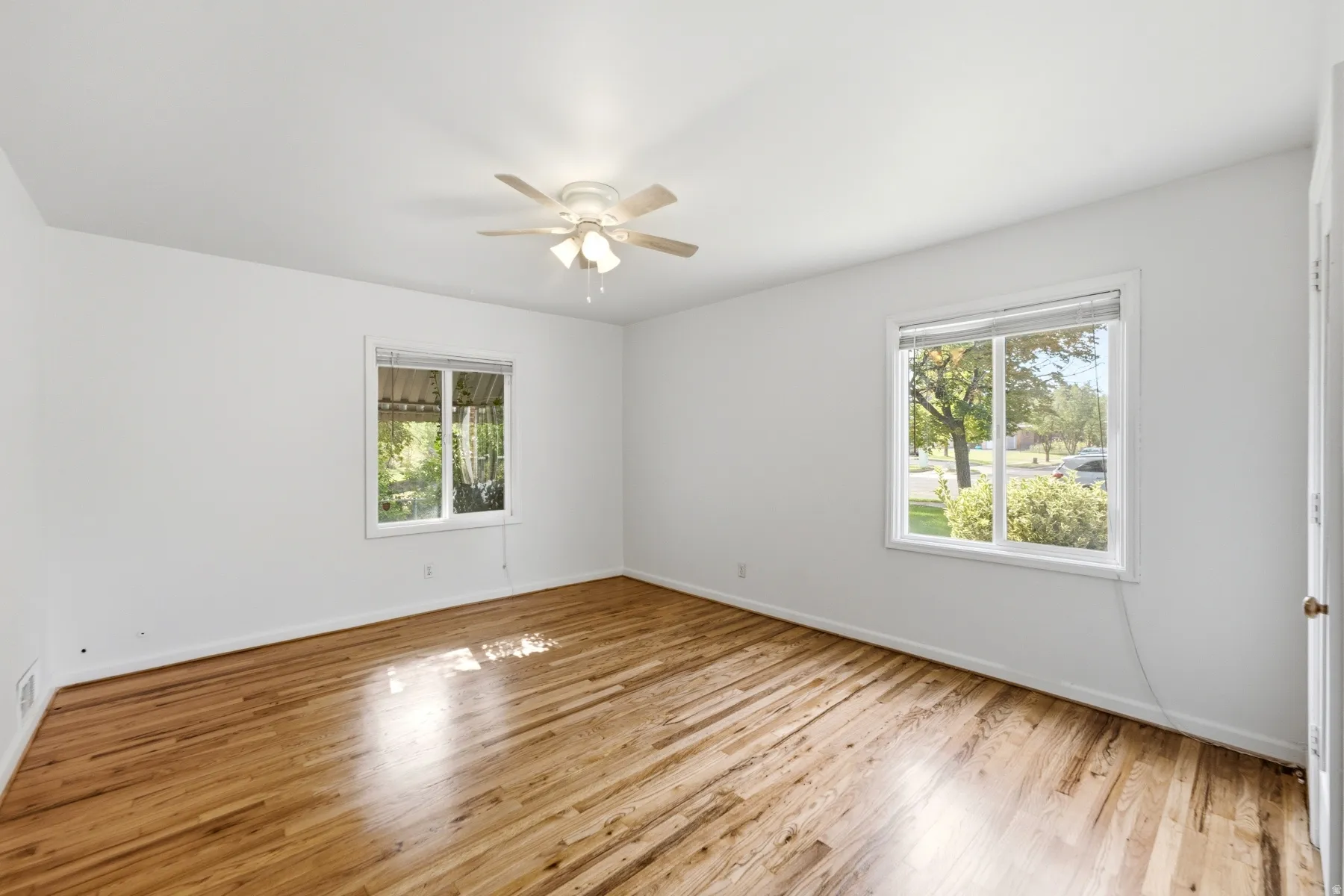 Bright room with wood-finish flooring, two large windows, a ceiling fan with integrated lighting, and white walls