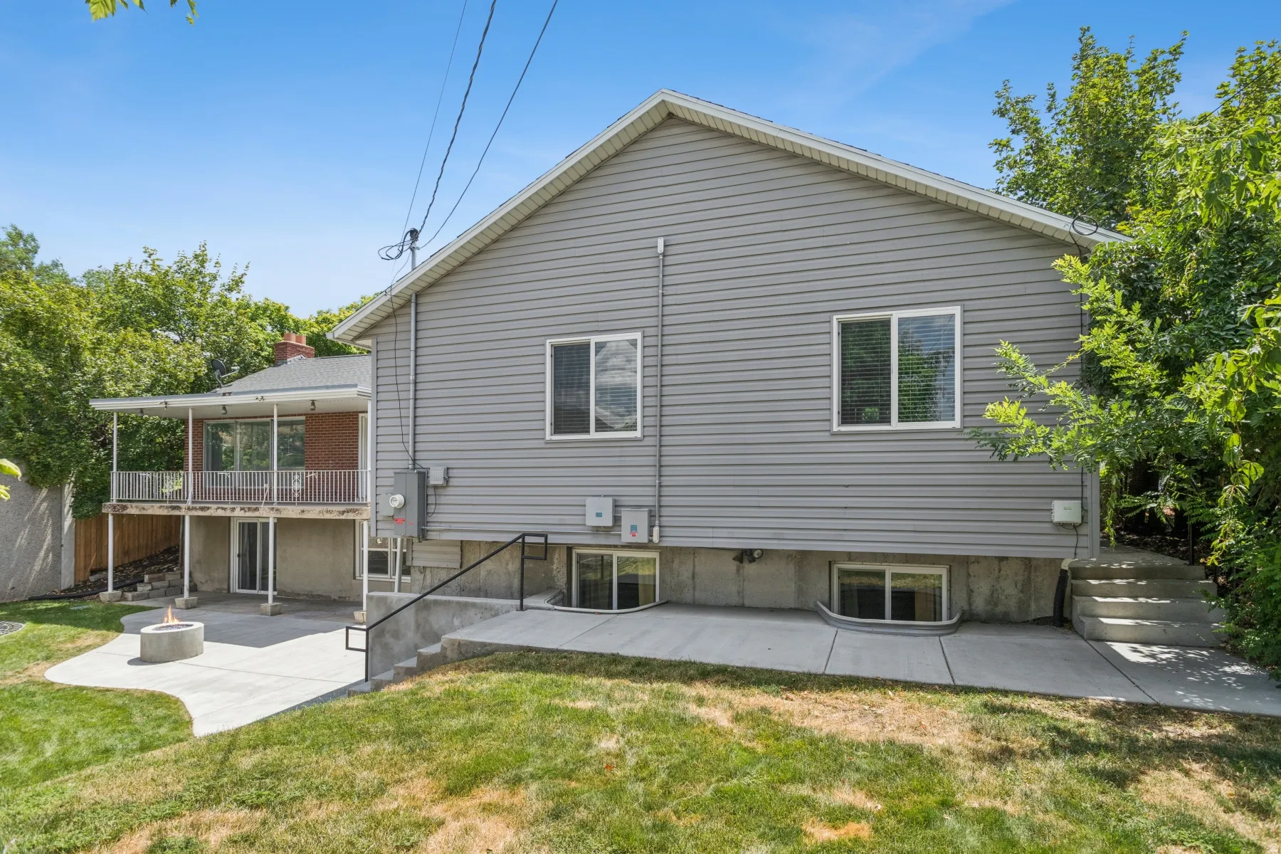 Gray siding exterior with a covered balcony, lower-level patio with a built-in fire pit, multiple walk-out doors, and a concrete staircase