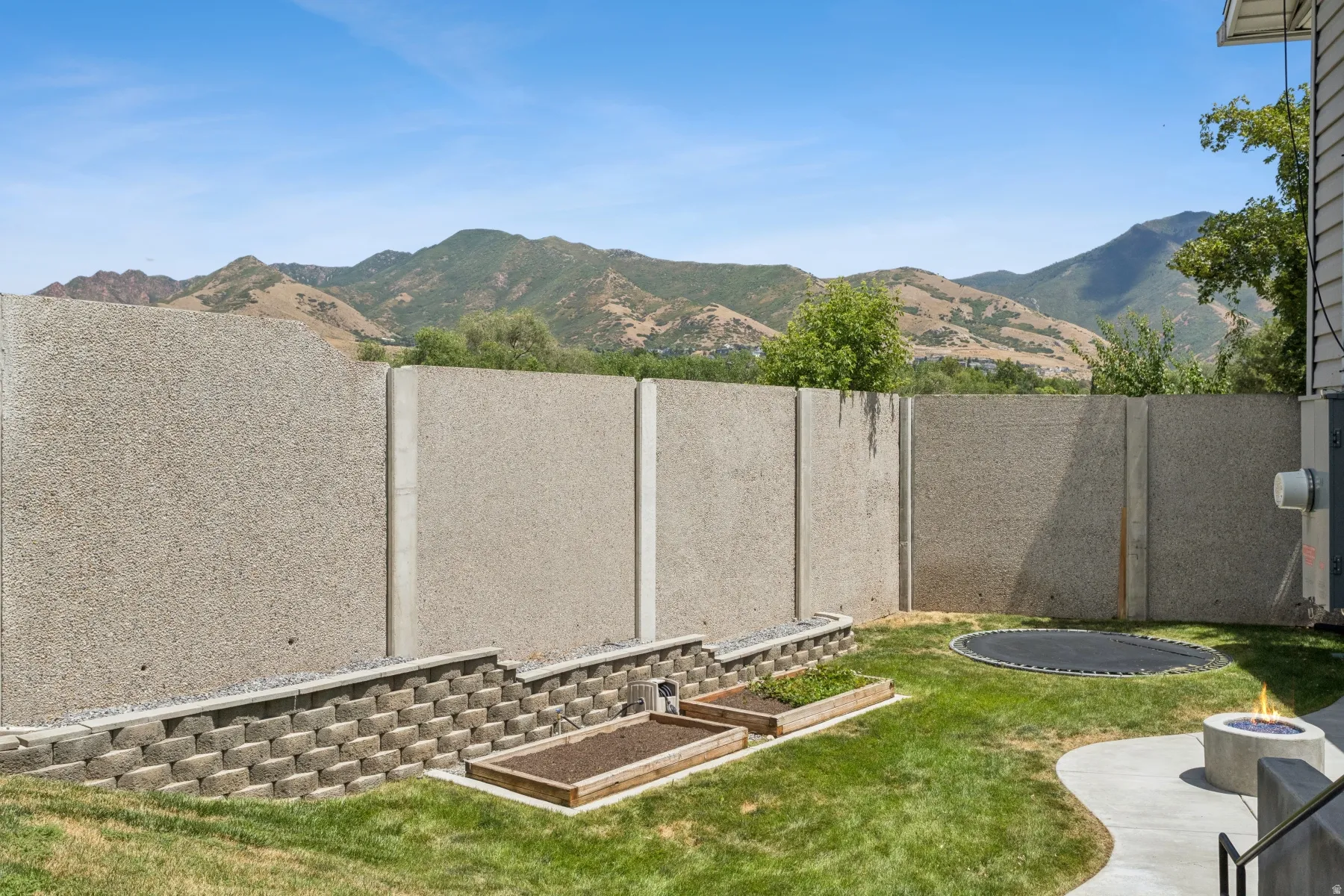 Expansive backyard featuring a concrete patio, built-in fire pit, and terraced garden beds with a retaining wall