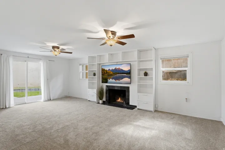Spacious room featuring a built-in entertainment center, fireplace with dark surround, sliding glass doors, multiple windows, and two ceiling fans