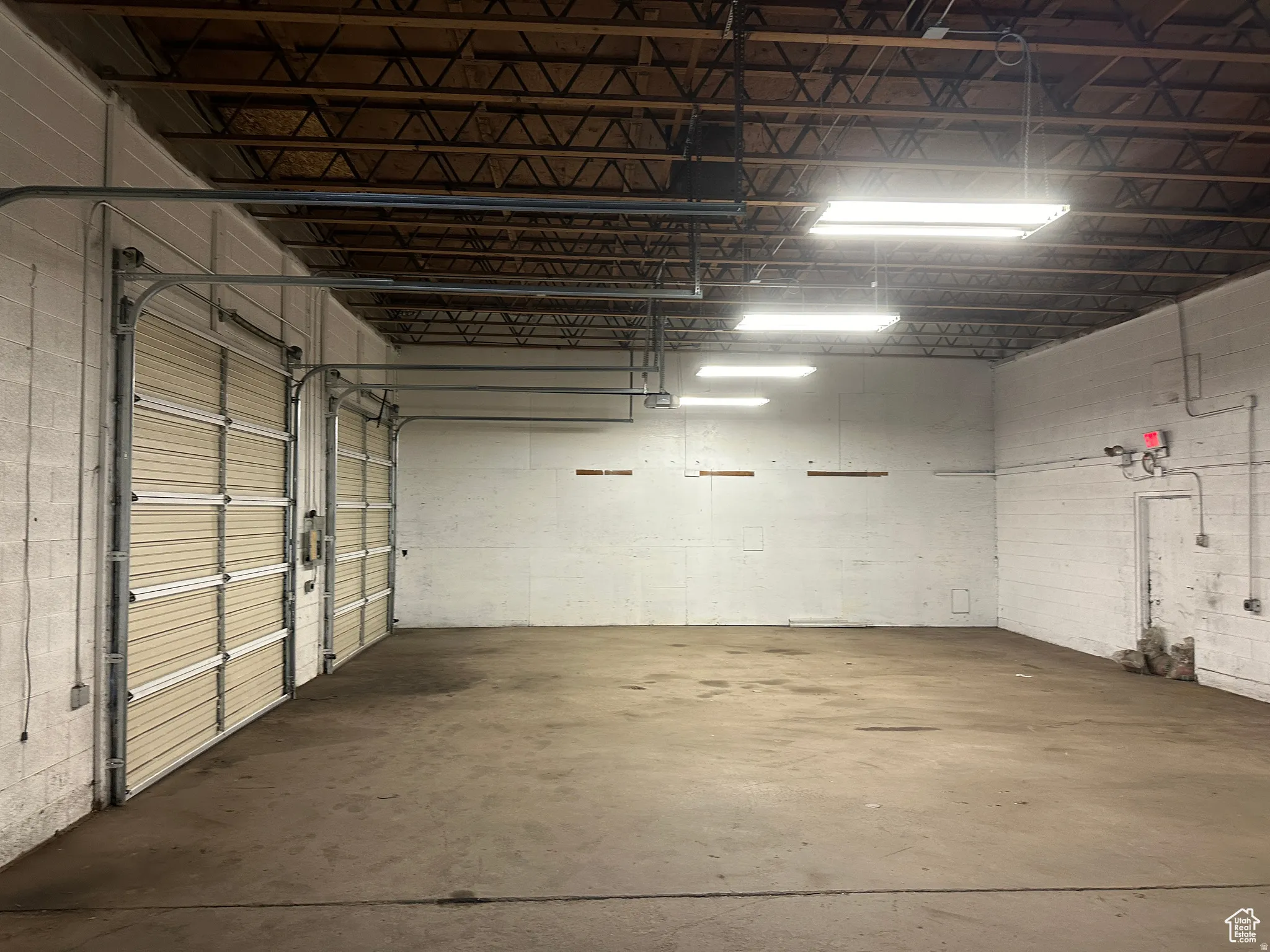 Concrete flooring, exposed ceiling trusses, multiple overhead fluorescent light fixtures, two rollup doors, and painted block walls