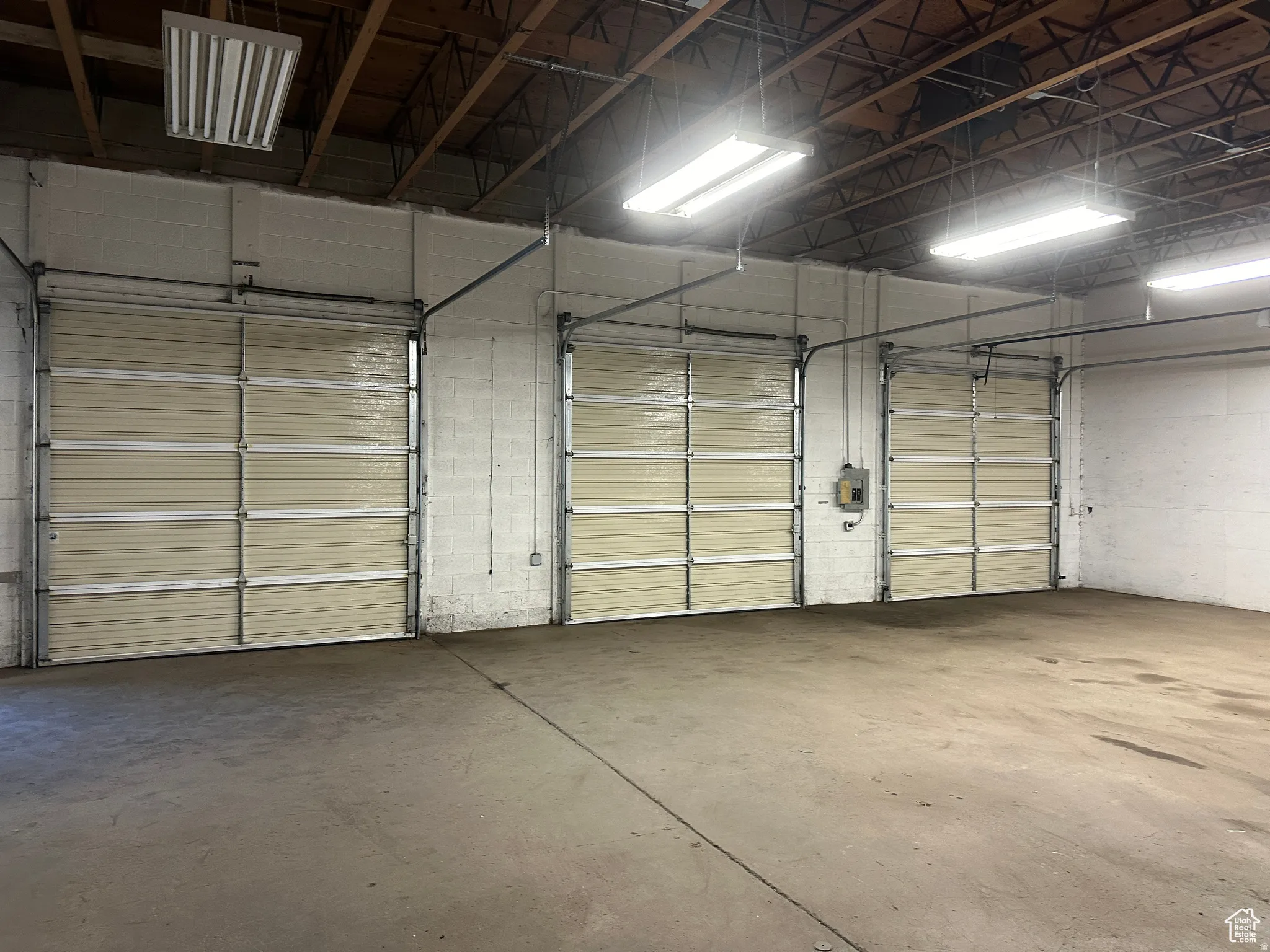 Three large roll-up doors, exposed ceiling trusses, concrete flooring, and suspended fluorescent lighting fixtures