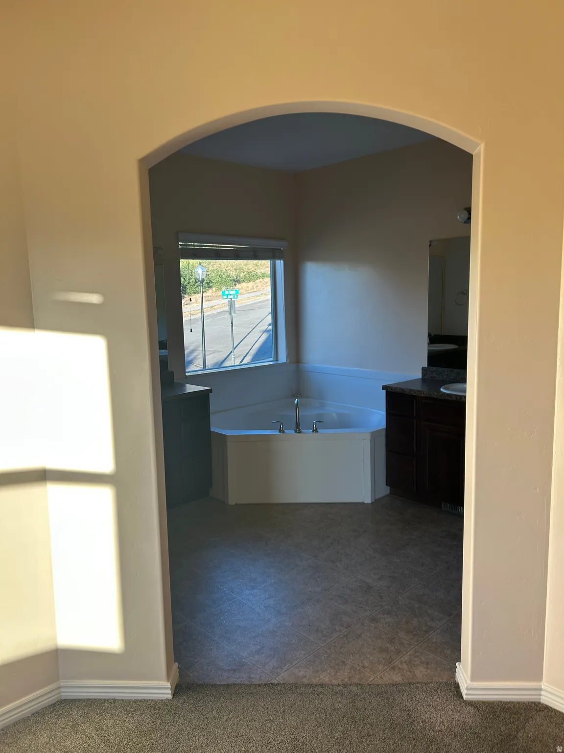 Arched doorway entry to a bathroom featuring a corner soaking tub, dual vanities with dark wood-finish cabinetry, and a window with blinds