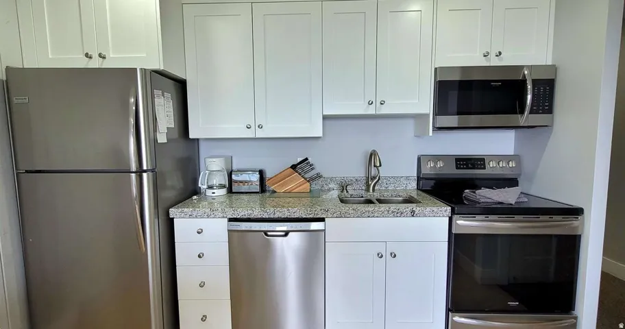 Updated kitchen featuring stainless steel appliances including a dishwasher, electric range, and refrigerator; white cabinetry with hardware accents; speckled quartz countertops; and under-cabinet lighting